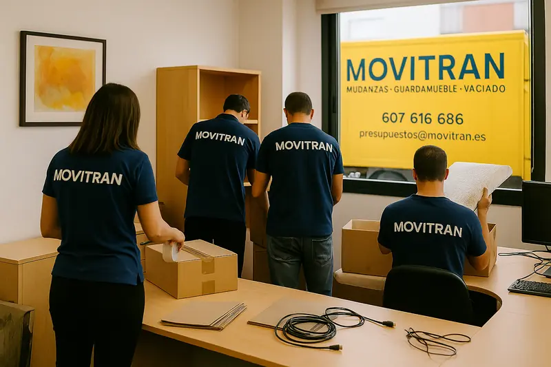 Equipo de Movitran embalando mobiliario y cajas en una oficina, con camión de mudanzas corporativo visible por la ventana.